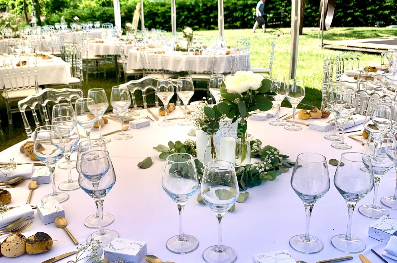 Table dressée pour un repas en extérieur sous une tente, avec vaisselle élégante, décorations florales et couverts dorés.