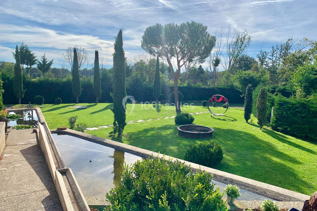 Jardin paysager avec pelouse, arbres, bassin d'eau et fauteuil suspendu sous un ciel partiellement nuageux.