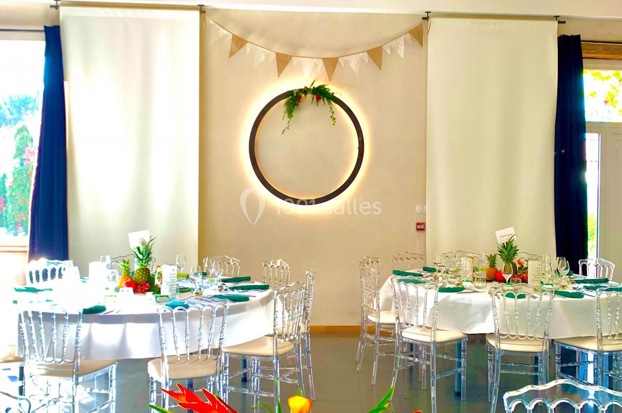 Salle de réception décorée avec des tables dressées, des fleurs tropicales et des guirlandes suspendues.