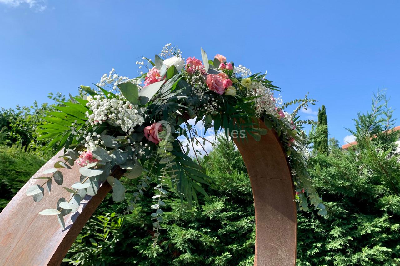 Arche en bois décorée de fleurs roses, blanches et de feuillage, installée dans un jardin sous un ciel bleu.