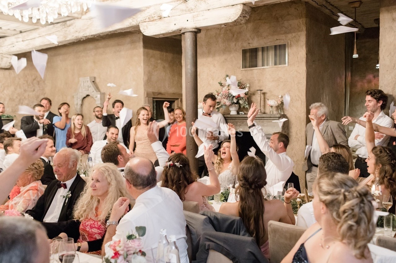 Des invités célèbrent joyeusement dans une salle de réception, lançant des avions en papier vers un couple au centre.