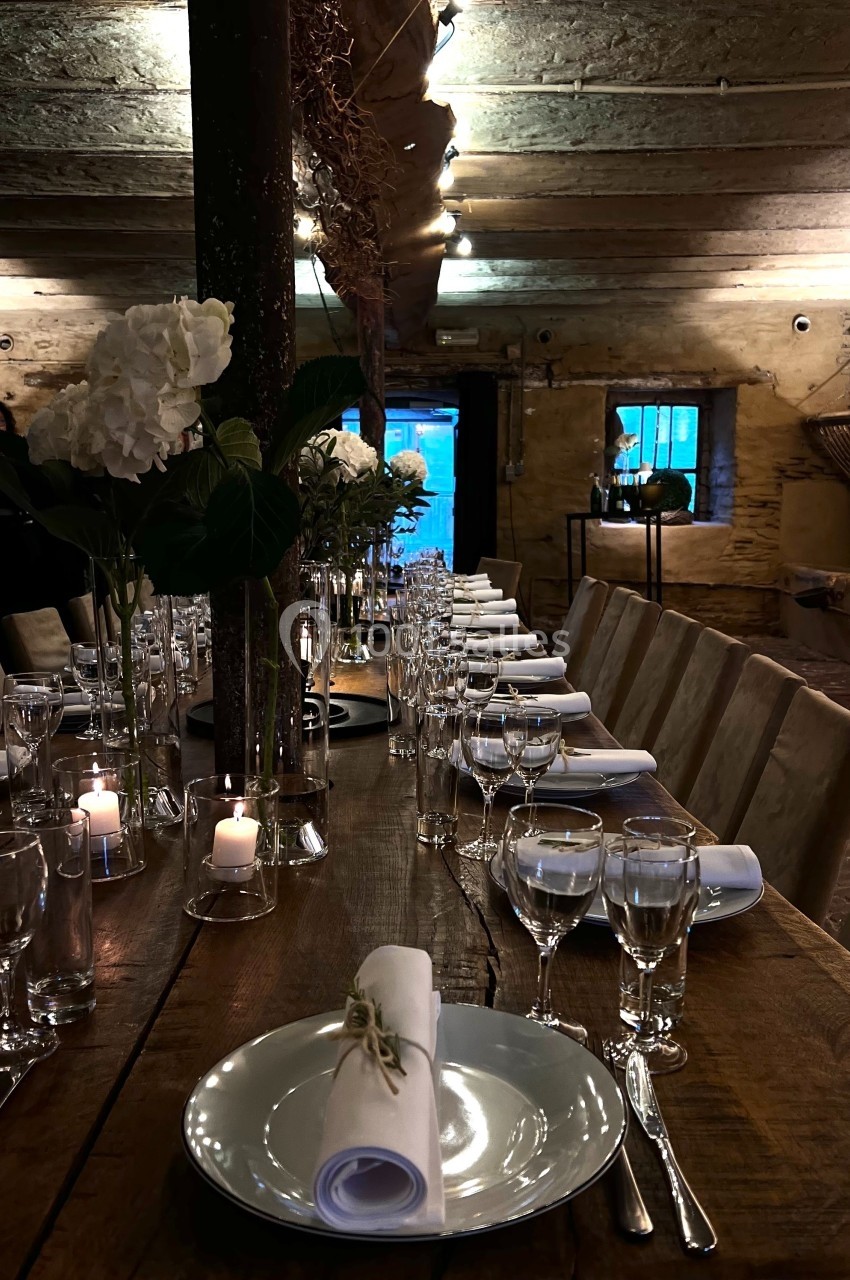 Table en bois décorée pour un dîner, avec vaisselle élégante, bougies et fleurs blanches dans un intérieur rustique.