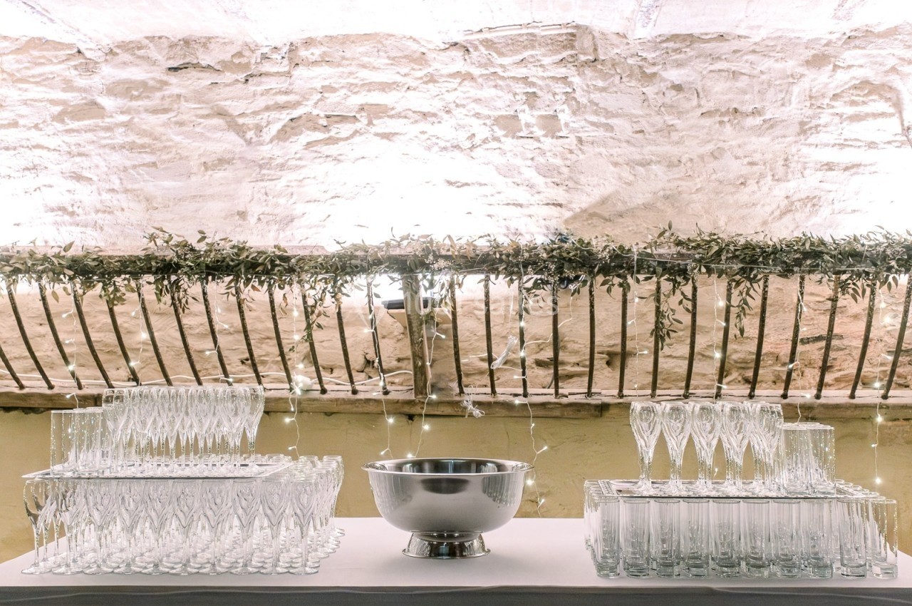 Table décorée avec des verres en cristal, une vasque en métal et une guirlande lumineuse sous un mur en pierre.