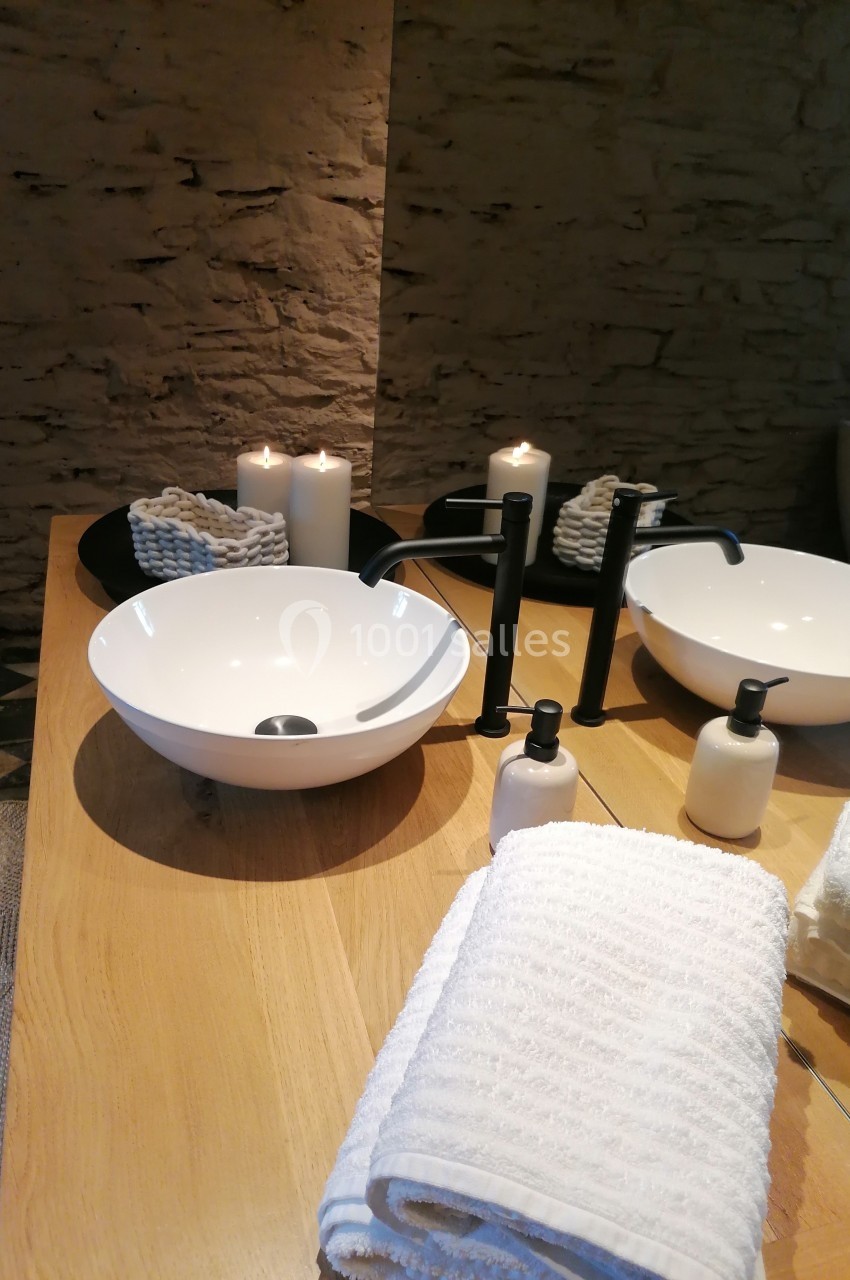 Deux vasques blanches sur un plan en bois, entourées de bougies, serviettes et flacons dans une salle de bain en pierre.