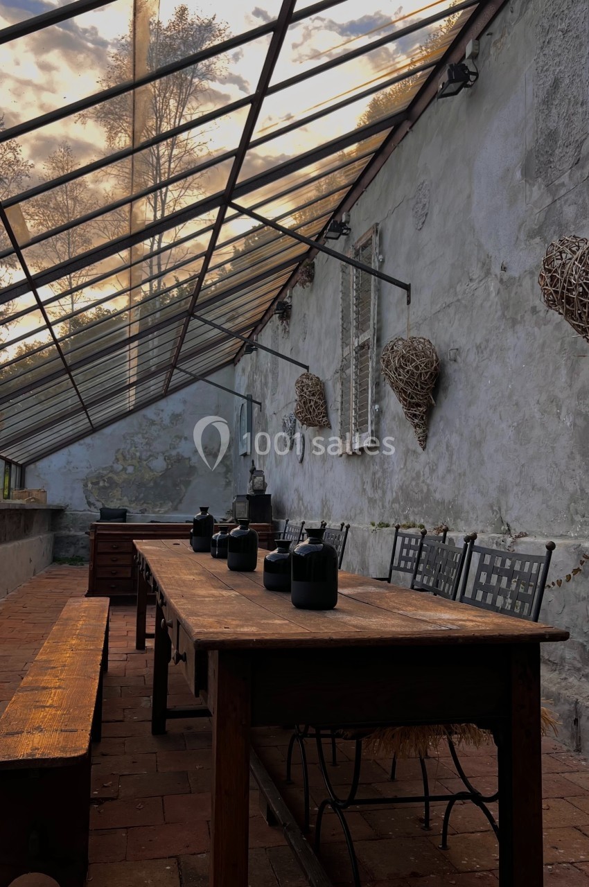 Table en bois avec des pots noirs, entourée de chaises, sous une verrière dans une cour au mur patiné.