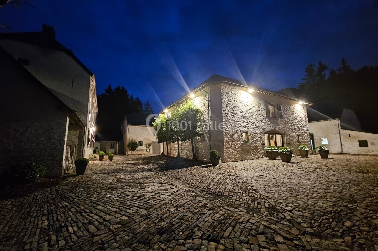 Cour pavée d'un bâtiment en pierre éclairé par des lumières extérieures, entouré d'arbres et de bâtiments annexes, de nuit.