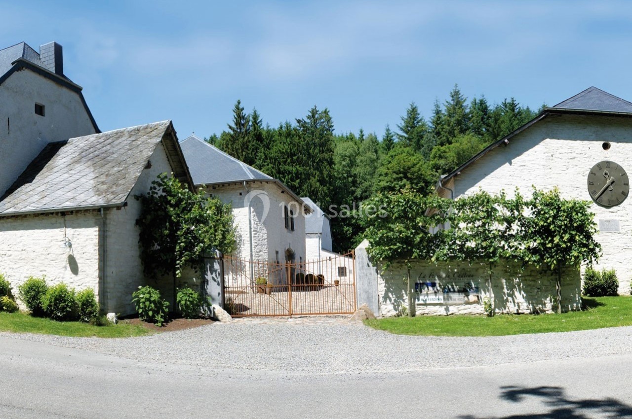 Portail en fer forgé entouré de bâtiments en pierre blanche avec toits en ardoise, bordés de végétation et d'arbres.