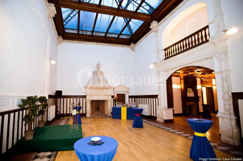 Salle lumineuse avec parquet, cheminée en pierre, tables hautes drapées de bleu et verrière en plafond.
