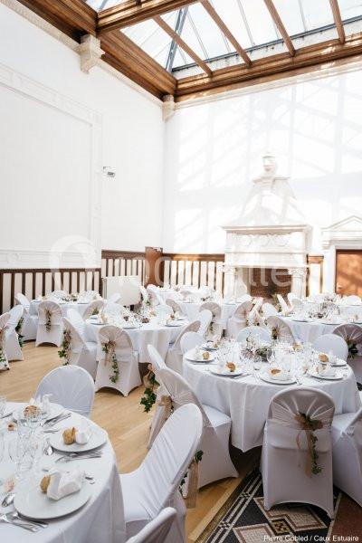 Salle lumineuse avec tables rondes dressées pour un événement, décorées de nappes blanches et de chaises ornées de rubans.