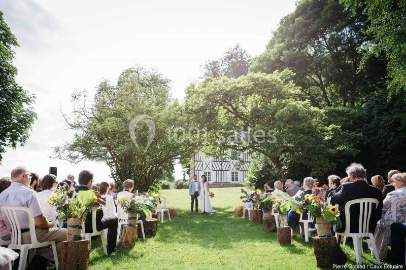 Cérémonie de mariage en plein air avec des invités assis, face à un couple devant une maison à colombages.