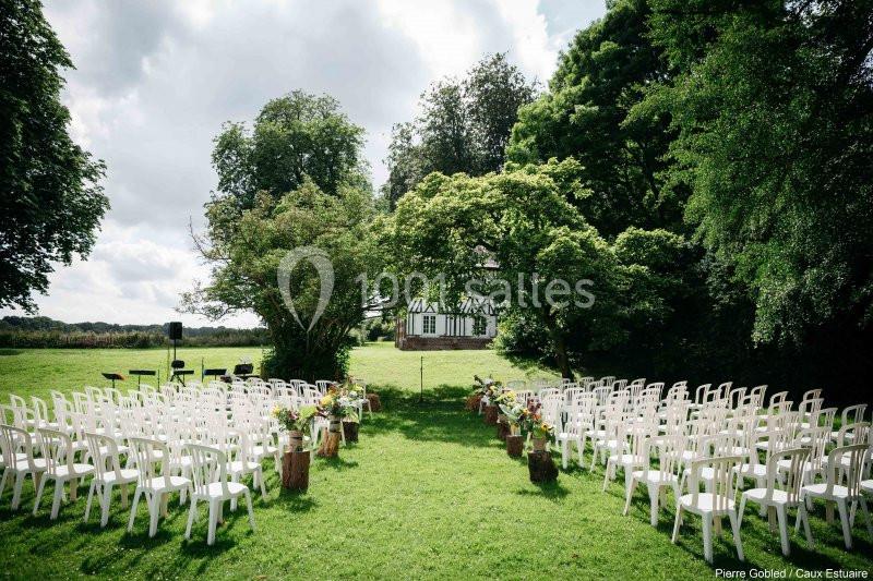 Chaises blanches disposées en rangées sur une pelouse, formant une allée menant à une petite maison à colombages.