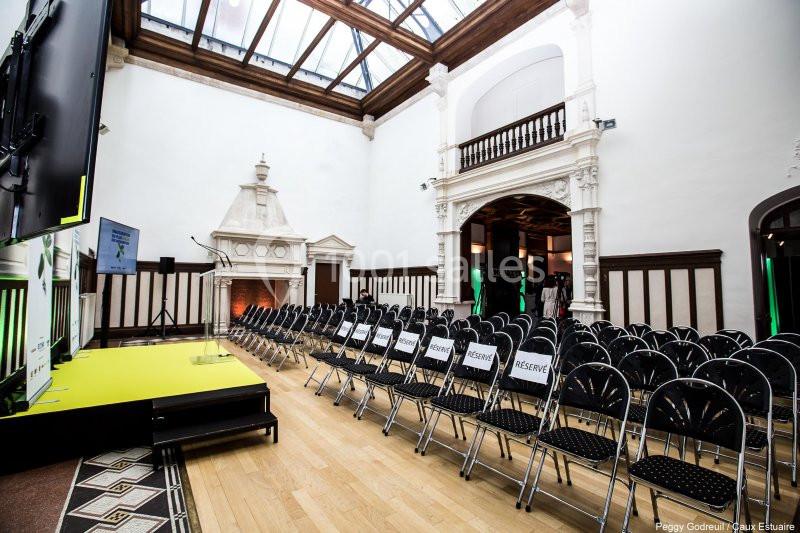 Salle lumineuse avec parquet, rangées de chaises noires alignées, scène jaune à gauche et cheminée décorative au fond.