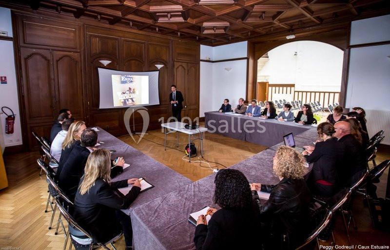Un groupe de personnes assises autour d'une table en U assiste à une présentation projetée dans une salle en bois.