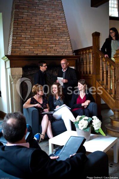 Un groupe de personnes échange dans un salon près d'une cheminée en briques, avec des documents et des fleurs blanches.