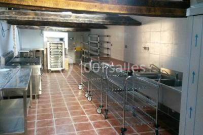 Cuisine professionnelle avec équipements en inox, plans de travail, éviers, chariots et plafond avec poutres apparentes.