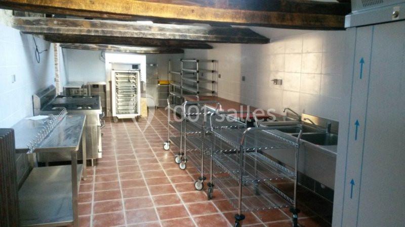 Cuisine professionnelle avec équipements en inox, plans de travail, éviers, chariots et plafond avec poutres apparentes.