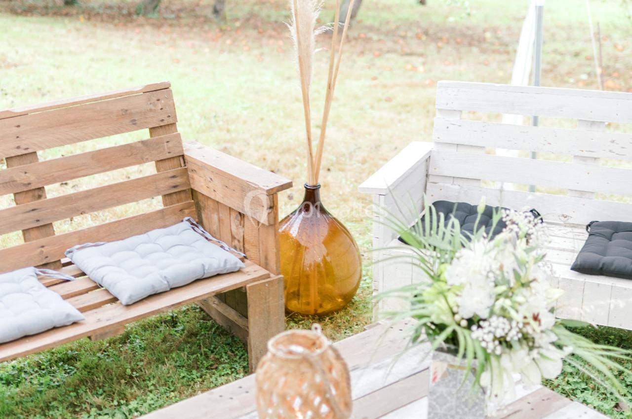 Salon de jardin en bois avec coussins, table basse décorée de fleurs et lanternes, installé dans un verger.