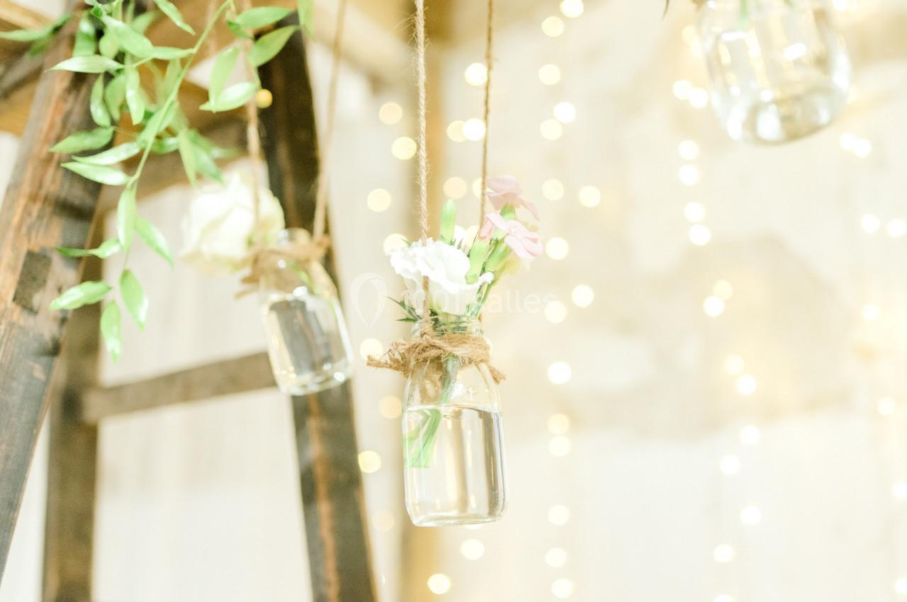 Petits vases suspendus en verre contenant des fleurs, accrochés à des cordes dans un décor lumineux et flou.