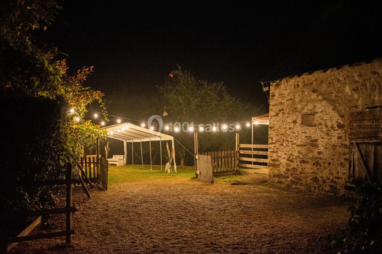 Cour éclairée par des guirlandes lumineuses, avec une tente blanche et un bâtiment en pierre sous un ciel nocturne.
