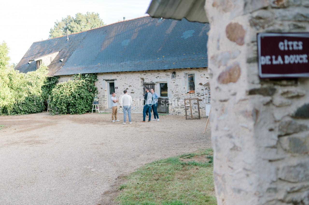 Un groupe de personnes discute devant un bâtiment en pierre avec un toit en ardoise, entouré de verdure.