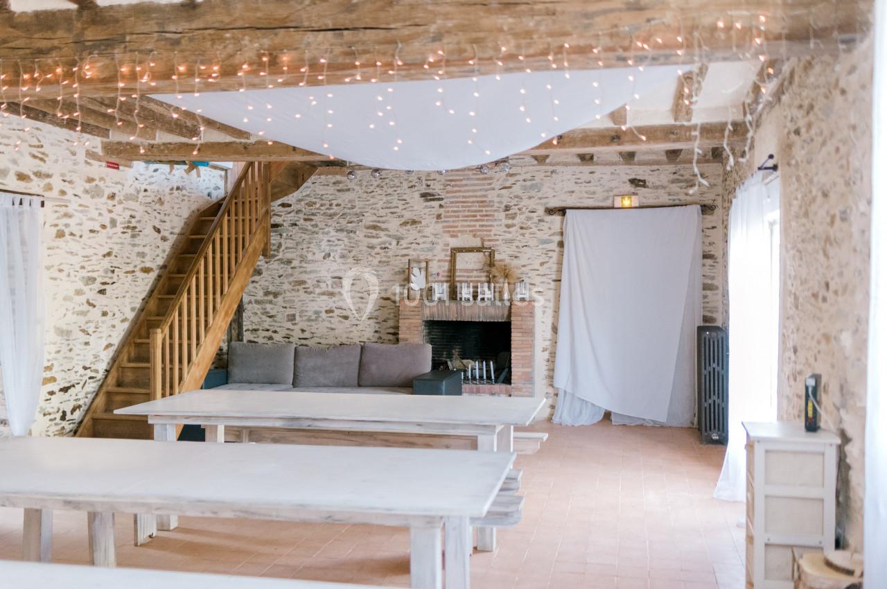 Salle lumineuse avec murs en pierre, poutres apparentes, cheminée, tables en bois clair et guirlandes lumineuses.