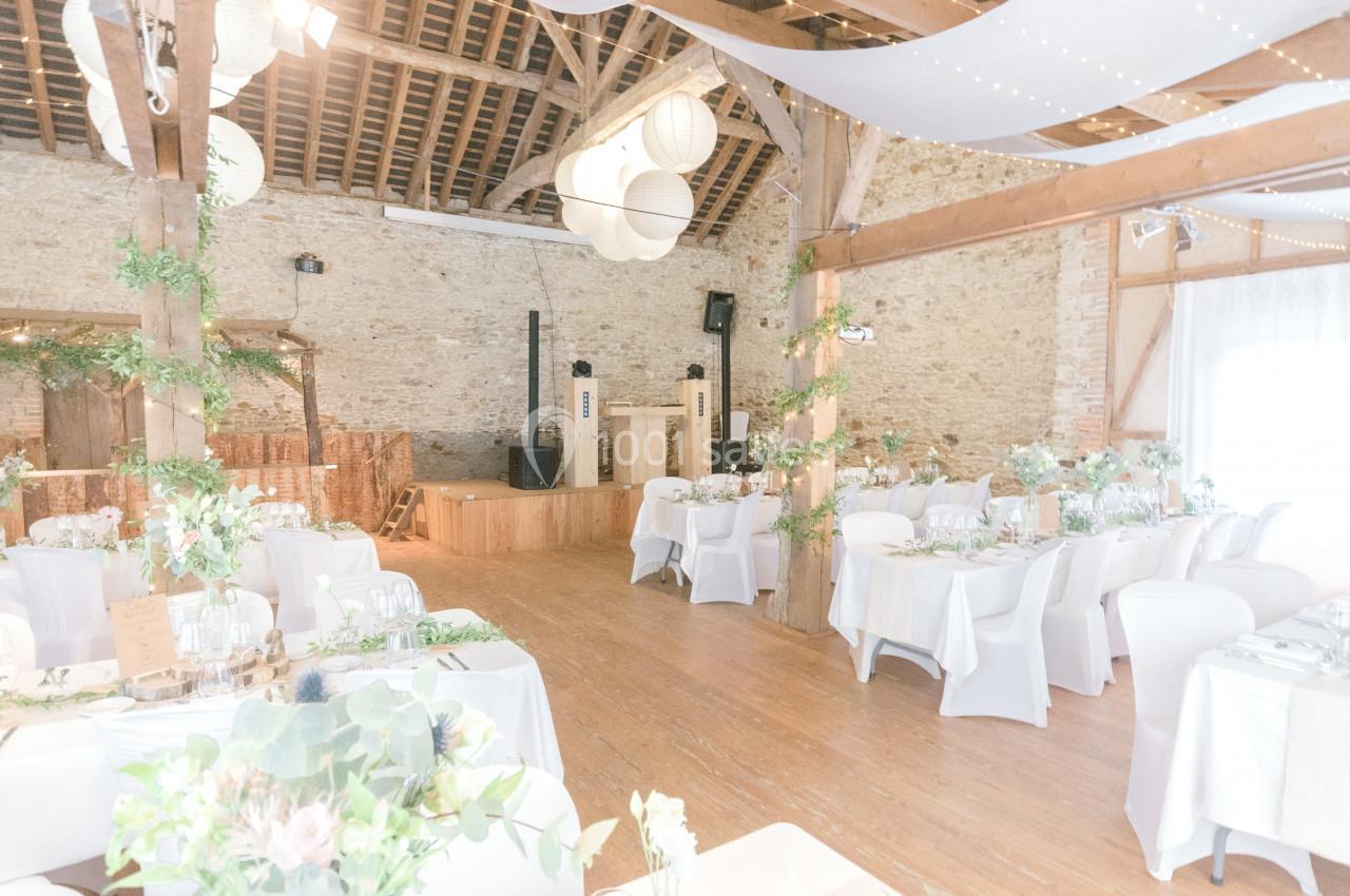 Salle de réception lumineuse décorée pour un mariage, avec tables dressées, chaises blanches et éléments végétaux.