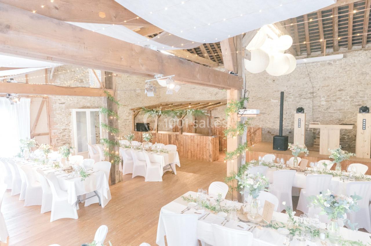 Salle de réception lumineuse avec tables décorées de fleurs, chaises blanches et guirlandes suspendues.