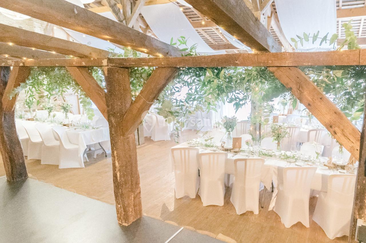 Salle de réception lumineuse avec tables décorées, chaises blanches et poutres en bois ornées de feuillage.
