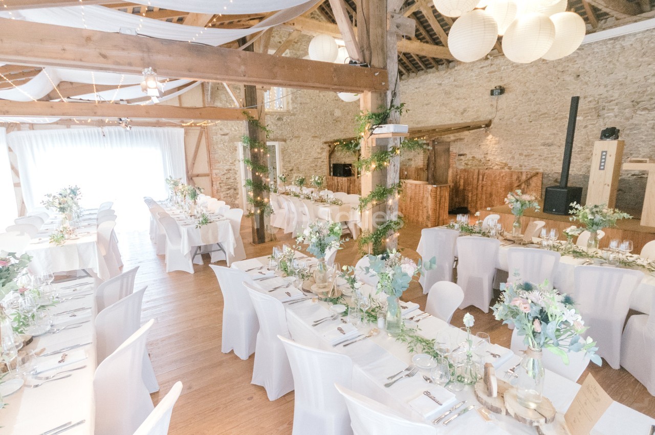 Salle de réception lumineuse décorée pour un mariage, avec tables dressées, chaises blanches et guirlandes végétales.