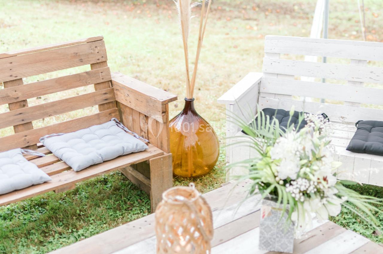 Bancs en bois avec coussins, vase en verre ambré et bouquet de fleurs blanches sur une table, dans un jardin.