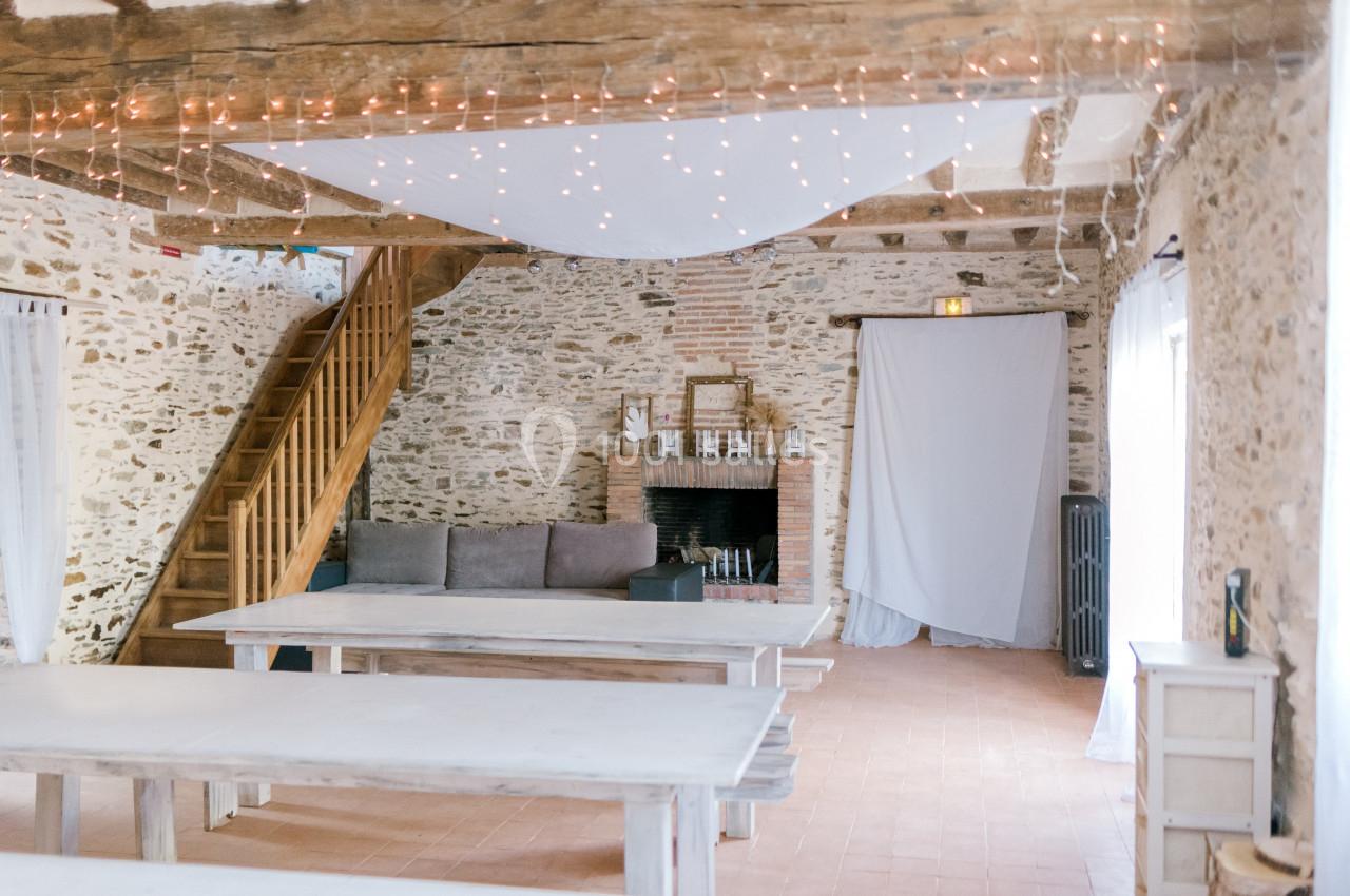 Salle lumineuse avec murs en pierre, poutres apparentes, tables en bois clair, canapé, cheminée et guirlandes lumineuses.
