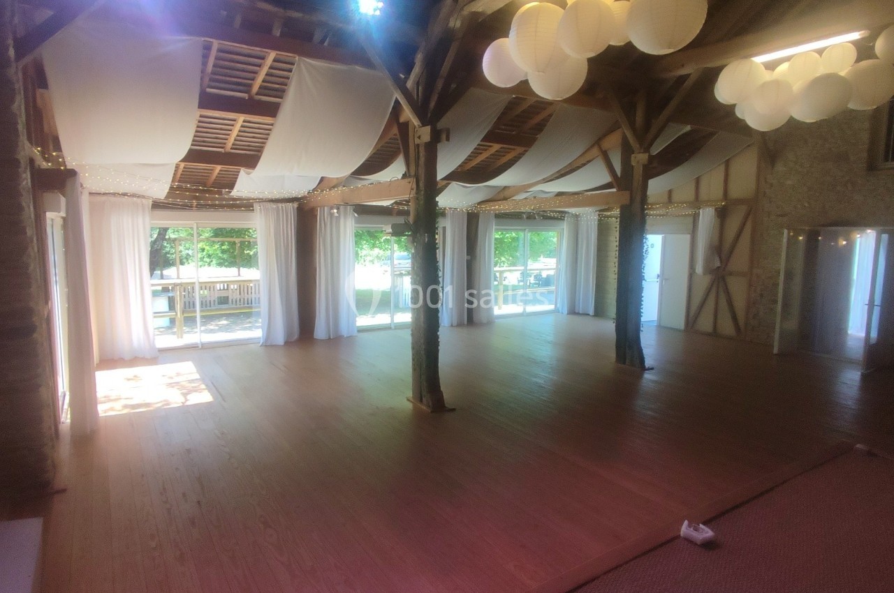 Salle lumineuse avec parquet en bois, grandes baies vitrées, poutres apparentes et décorations suspendues.