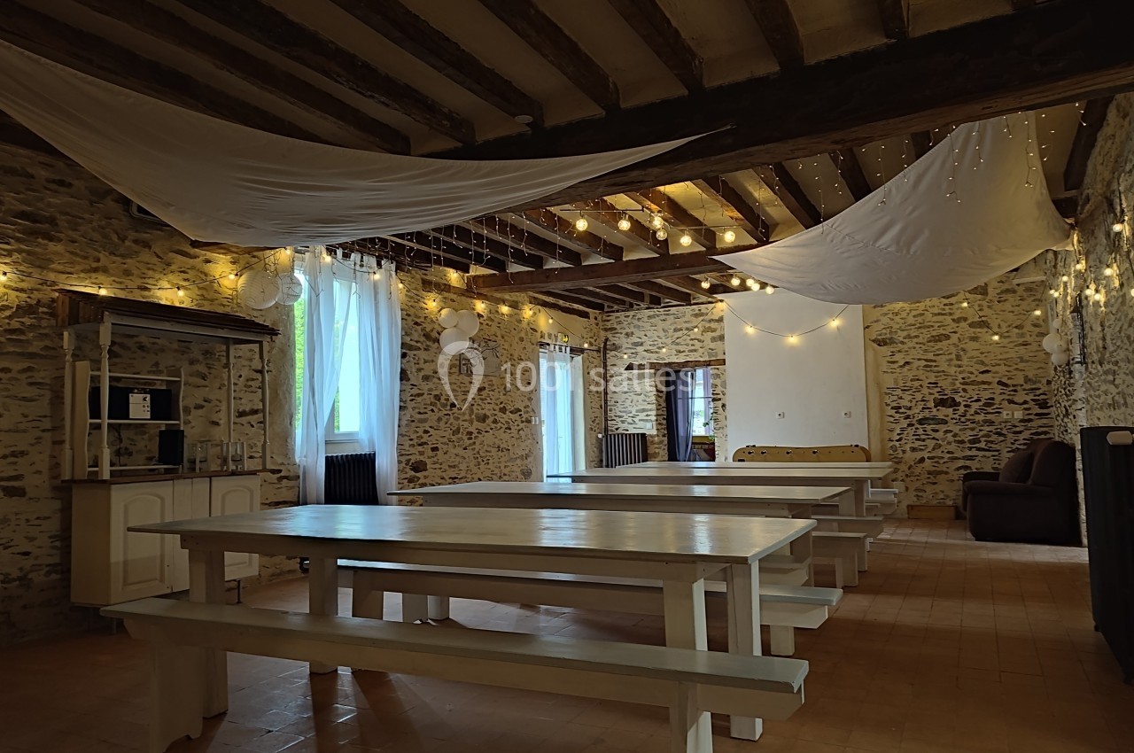 Salle rustique avec murs en pierre, grandes tables en bois, guirlandes lumineuses et voilages suspendus au plafond.