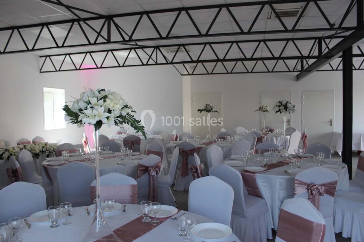 Salle de réception décorée avec des tables rondes, nappes blanches, chaises ornées de nœuds roses et centres de table…