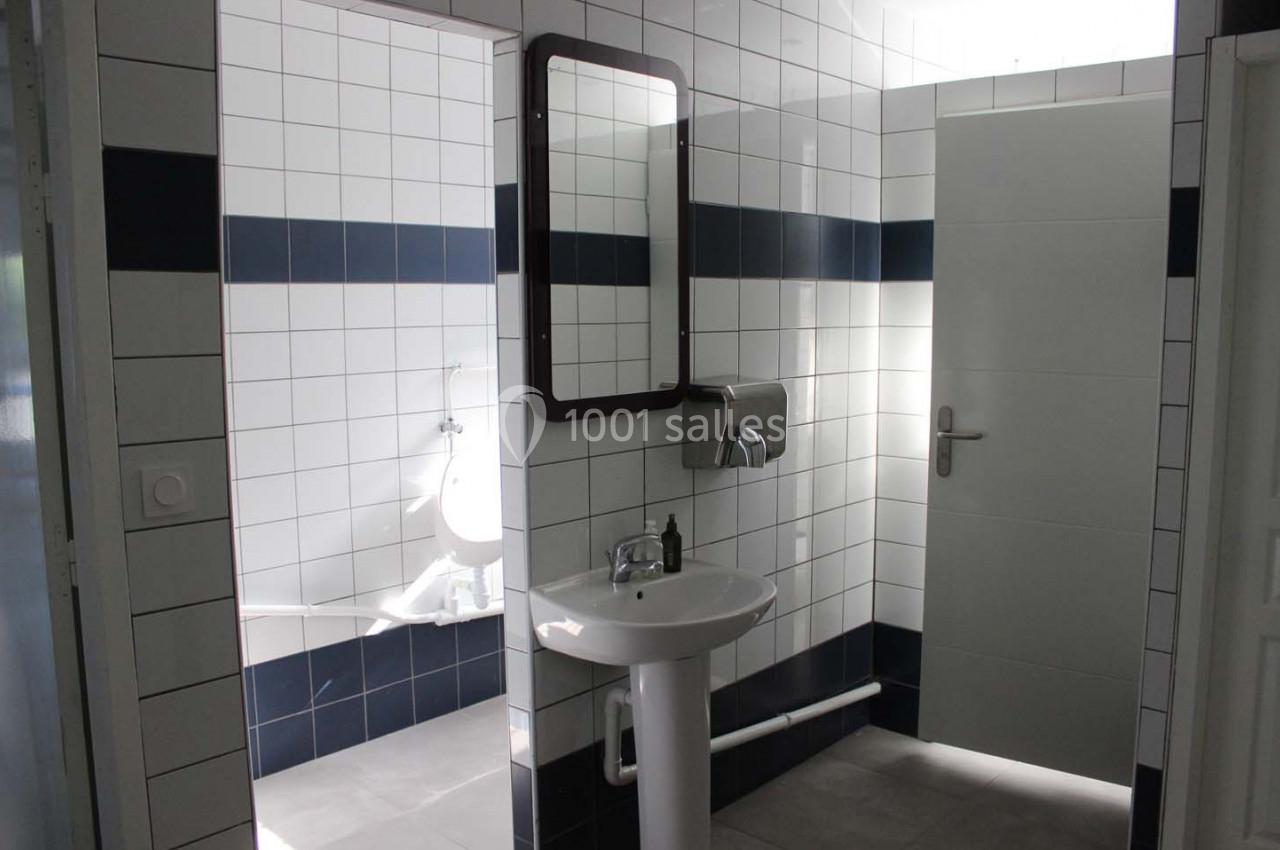 Salle de bain avec lavabo, miroir, urinoir et carrelage blanc et bleu, éclairée par une lumière naturelle.