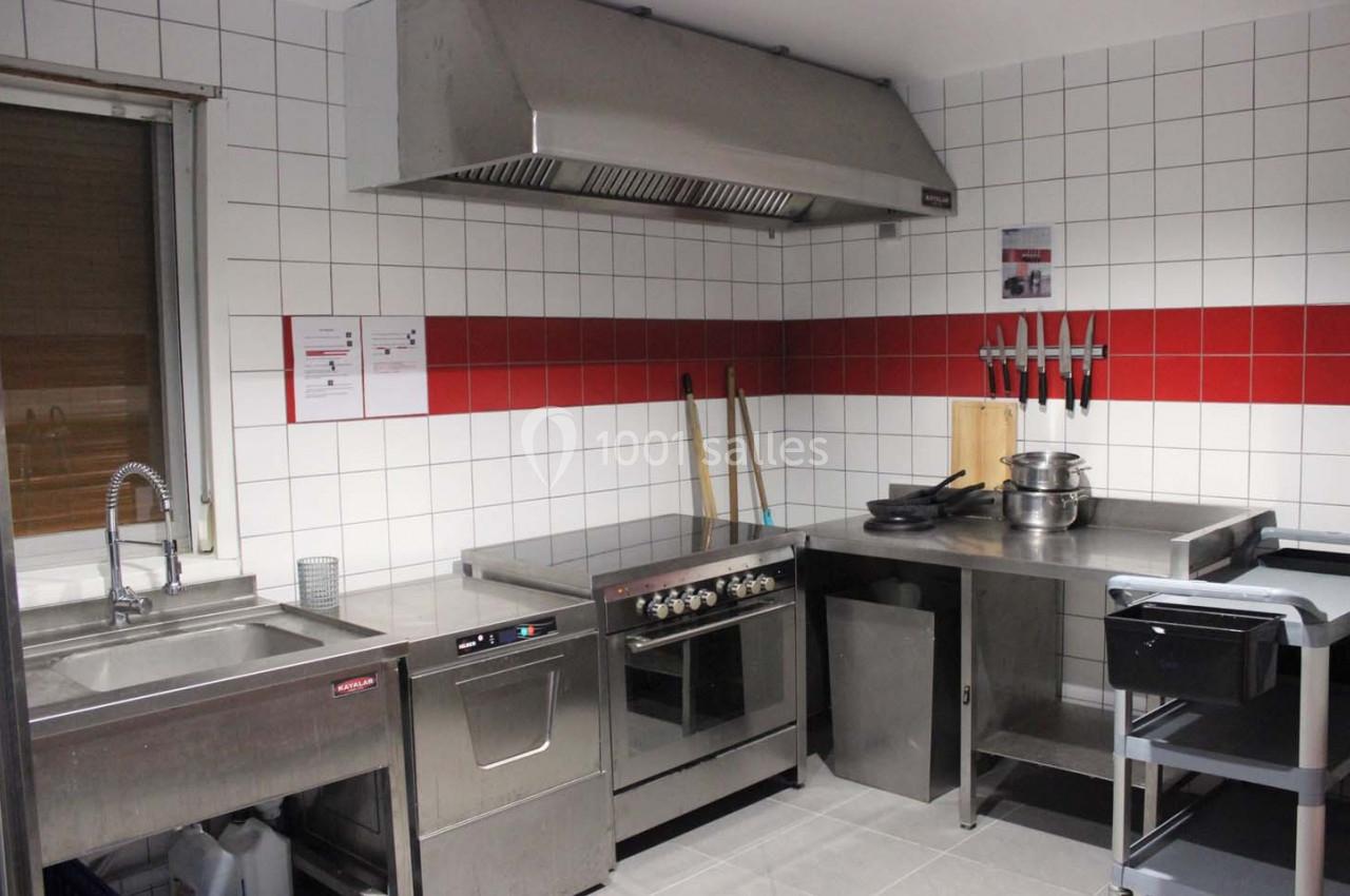 Cuisine professionnelle équipée avec évier, four, plaques de cuisson, hotte, plan de travail en inox et ustensiles accrochés.