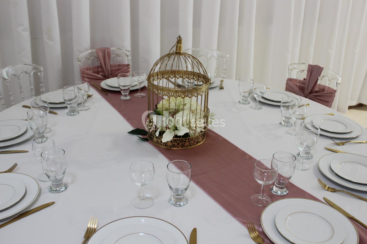 Table ronde dressée avec vaisselle blanche, couverts dorés, chemin de table rose et centre de table en cage dorée avec…