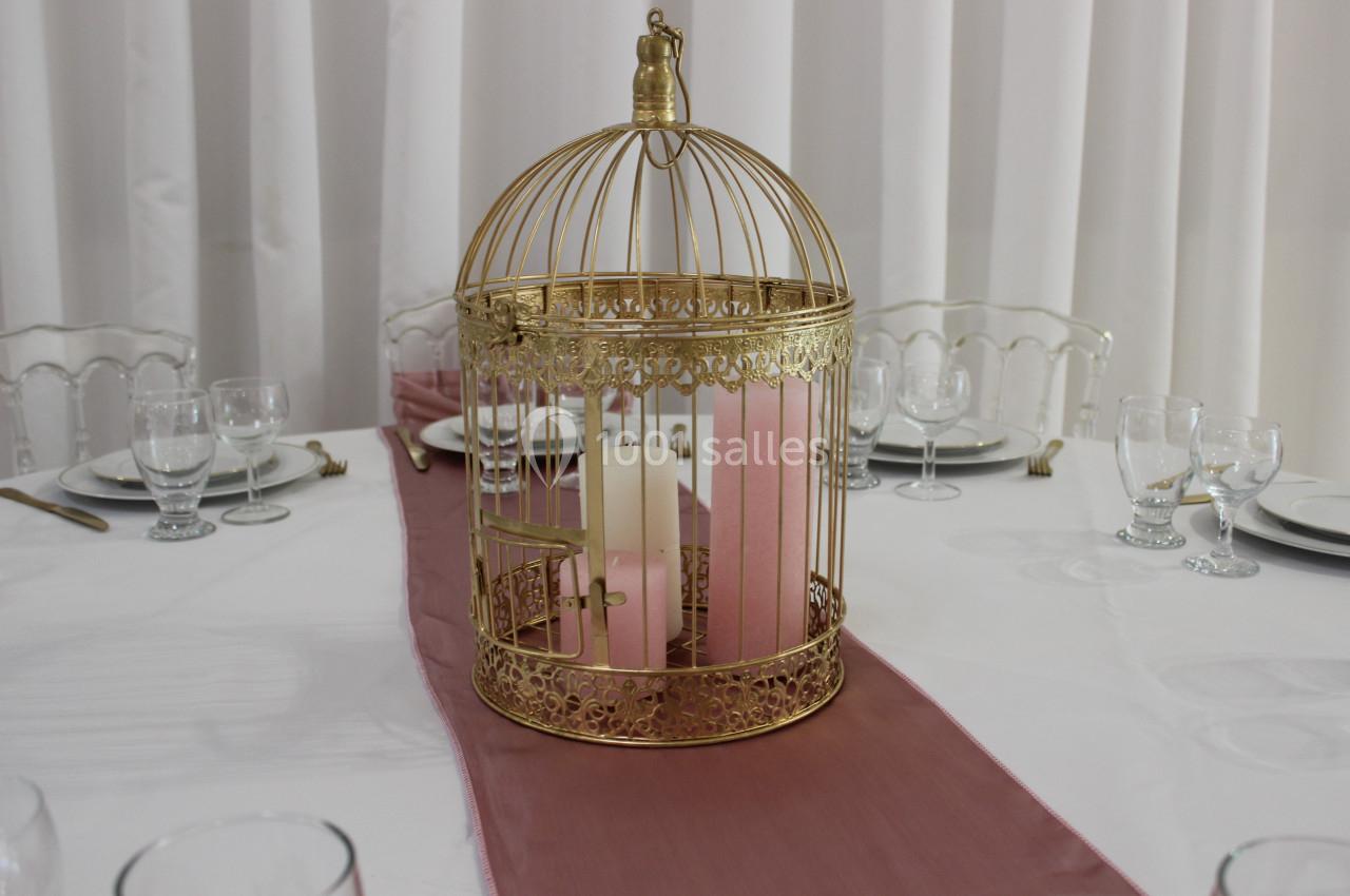 Centre de table avec une cage dorée contenant des bougies roses et blanches, posée sur un chemin de table rose.