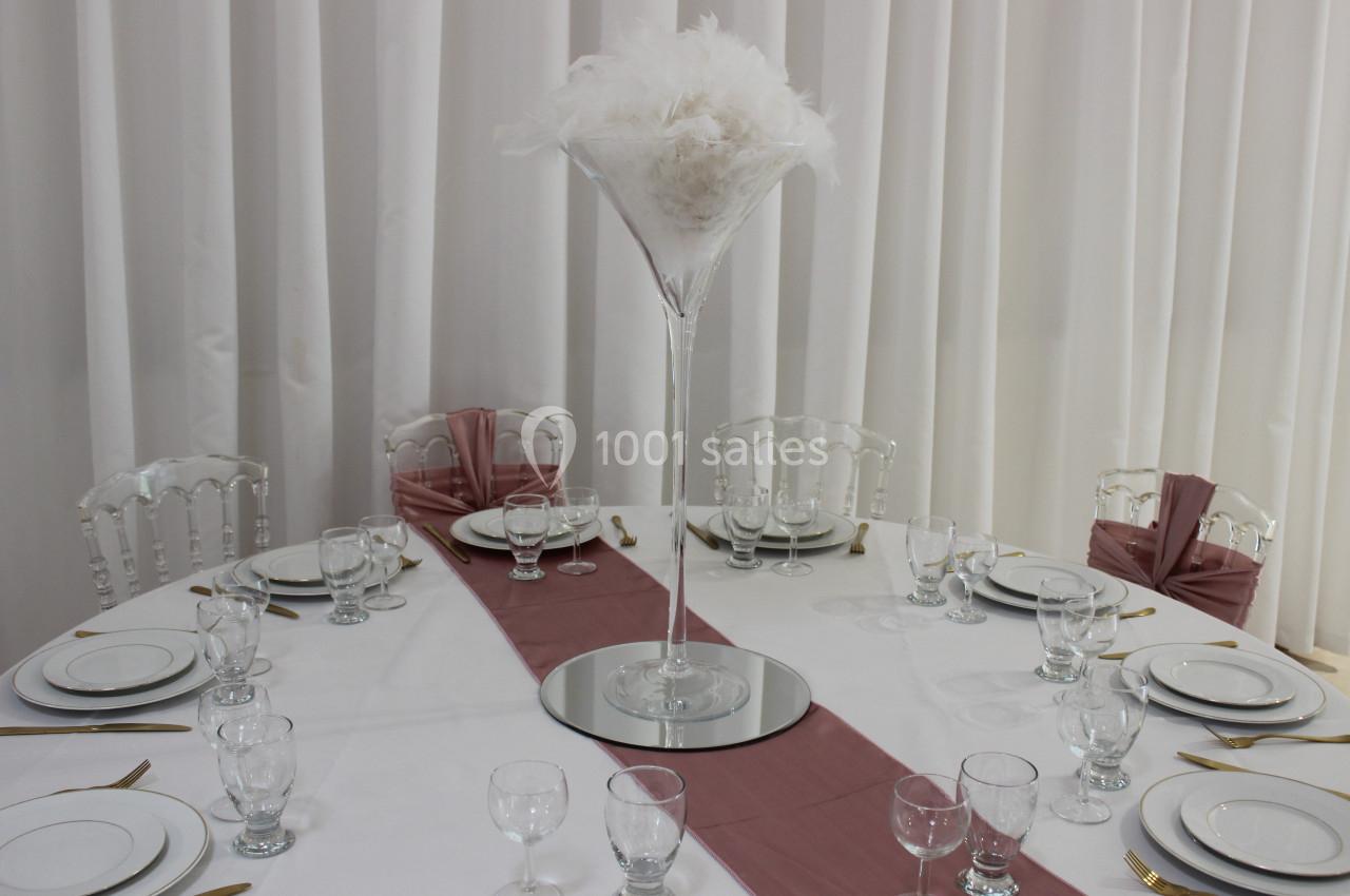 Table ronde dressée avec vaisselle blanche, couverts dorés, chemin de table rose et centre de table en plumes blanches.