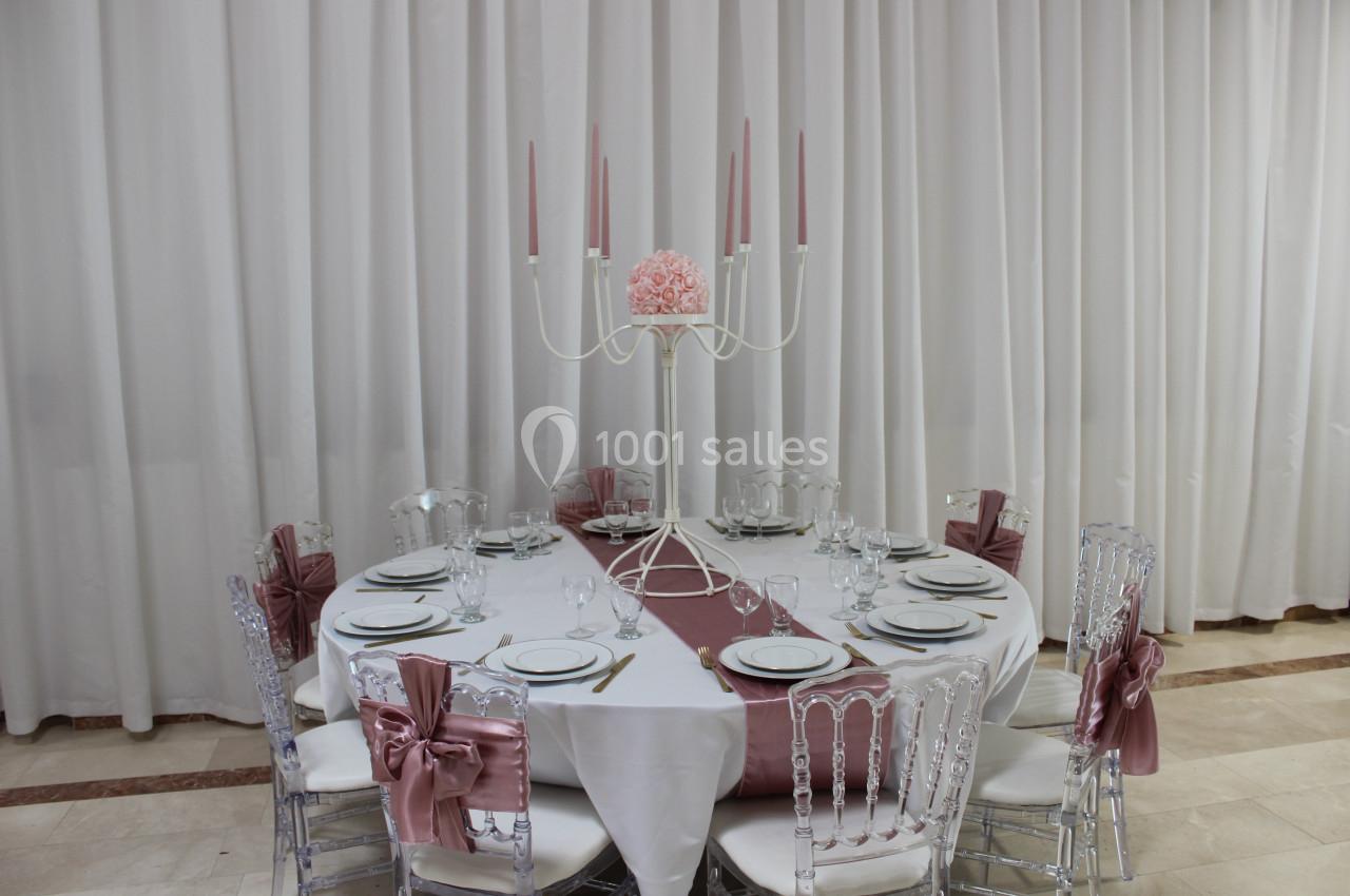 Table ronde élégamment dressée avec nappes blanches, chaises transparentes ornées de nœuds roses et chandelier central.