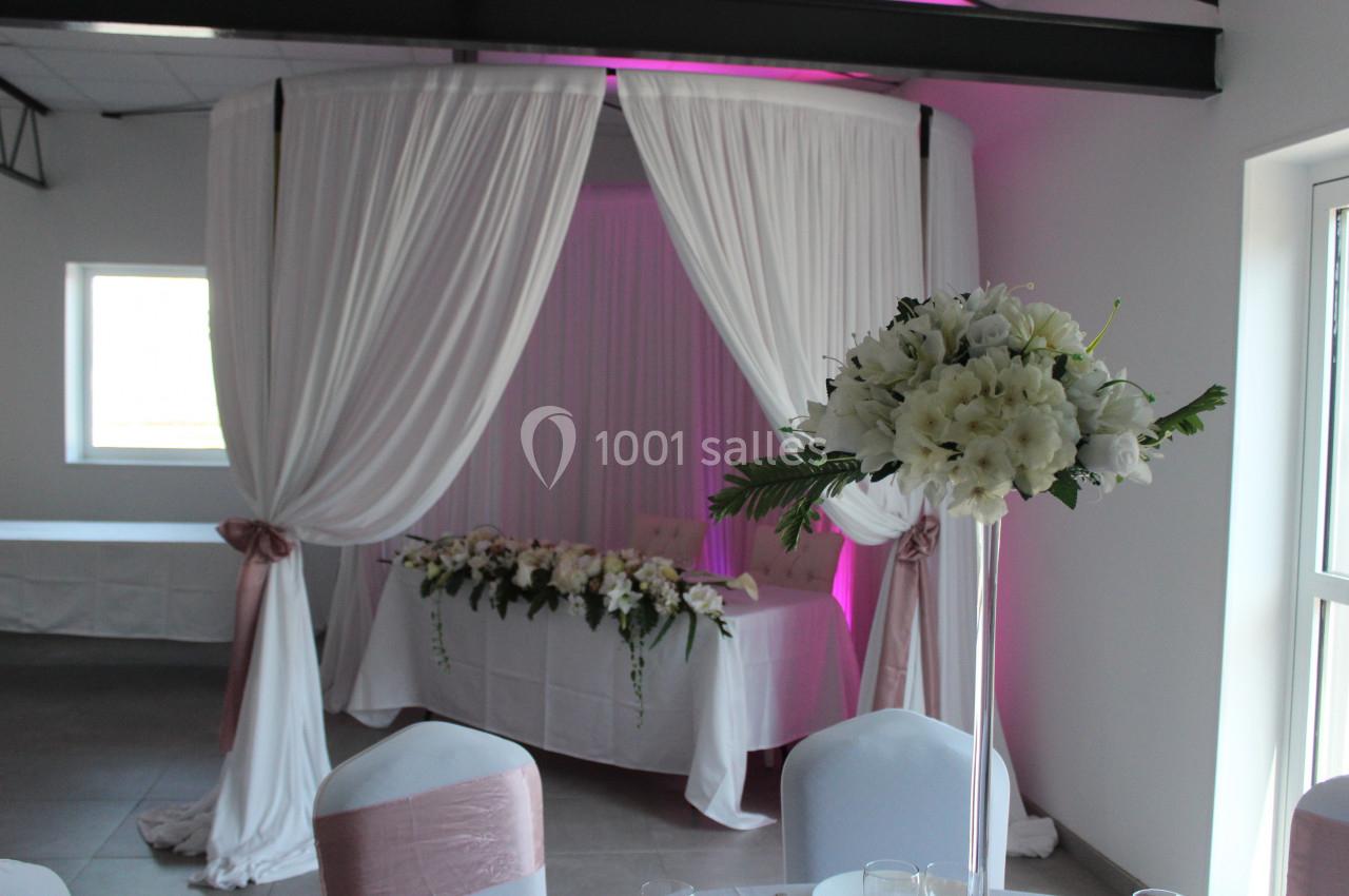 Décoration de salle avec arche drapée blanche, table ornée de fleurs et chaise habillée, éclairage rose en arrière-plan.