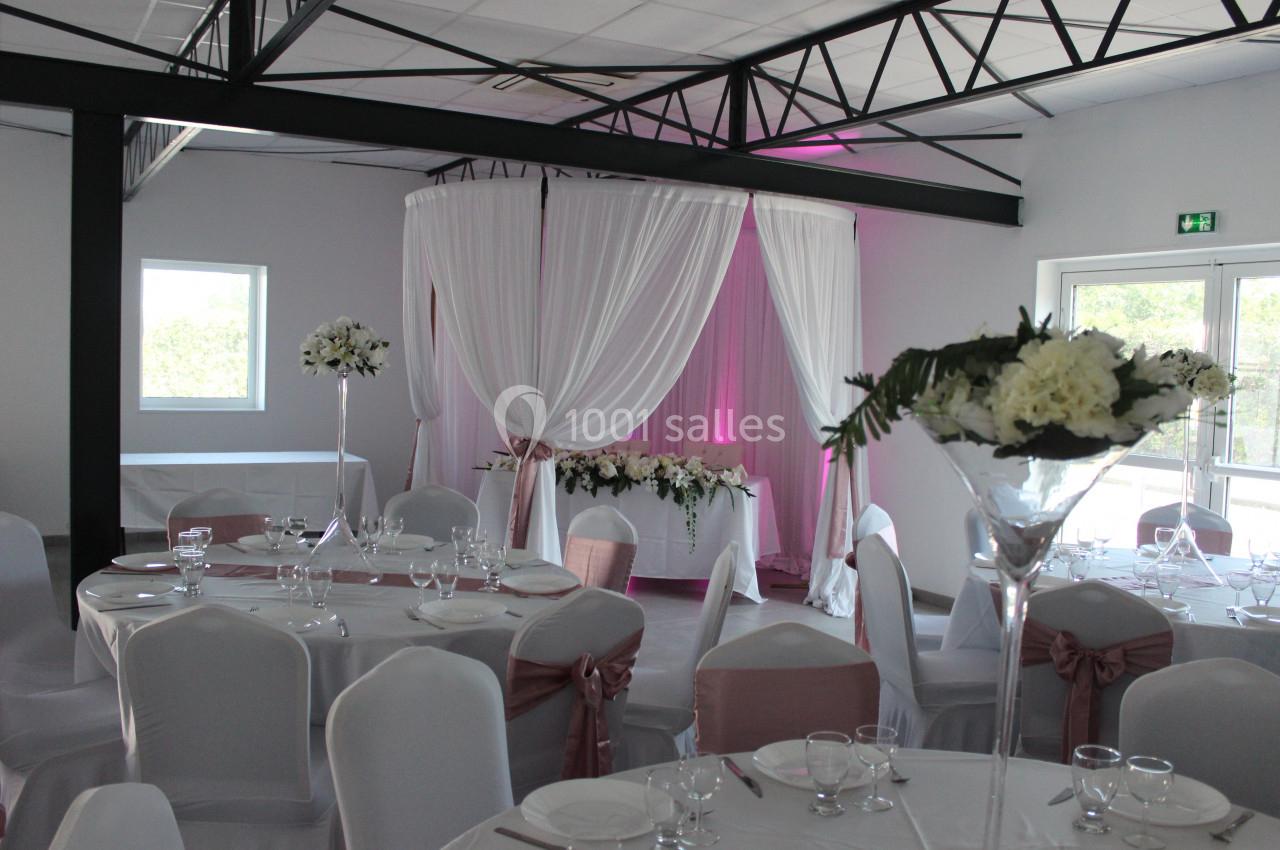 Salle de réception décorée avec des tables rondes, nappes blanches, chaises ornées et un espace avec rideaux blancs.