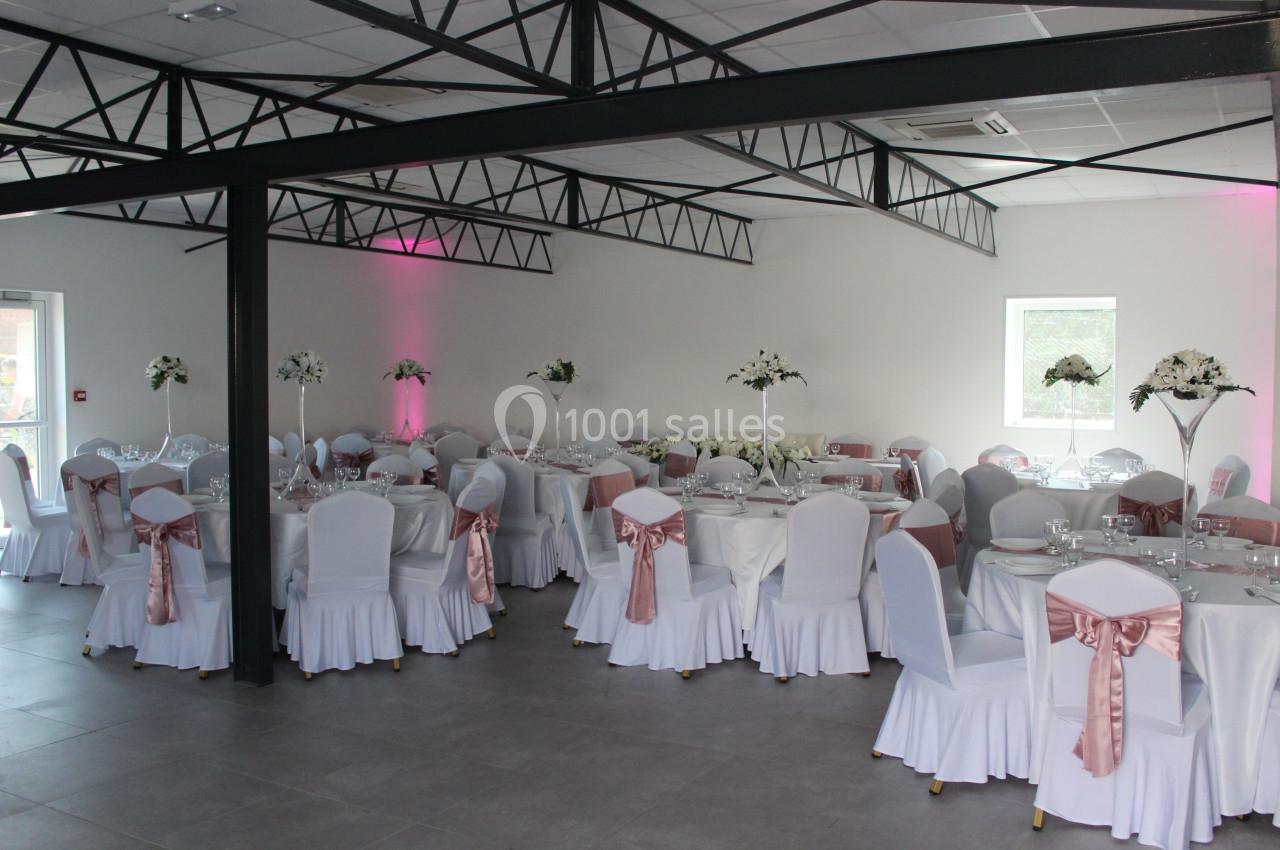 Salle de réception décorée avec des tables rondes, nappes blanches, chaises ornées de nœuds roses et centres de table…