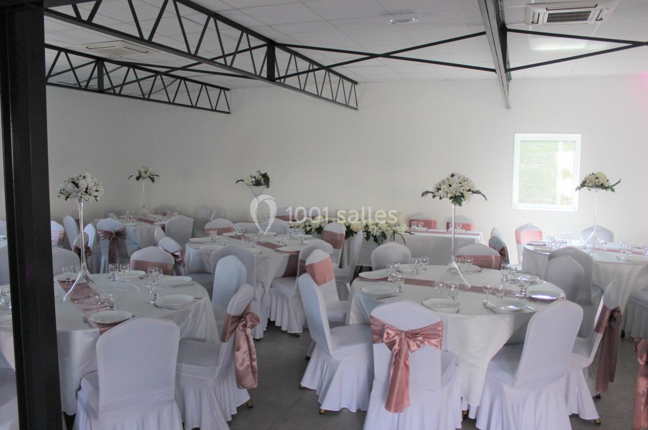 Salle décorée pour un événement, avec des tables rondes, nappes blanches, chaises ornées de rubans roses et compositions…