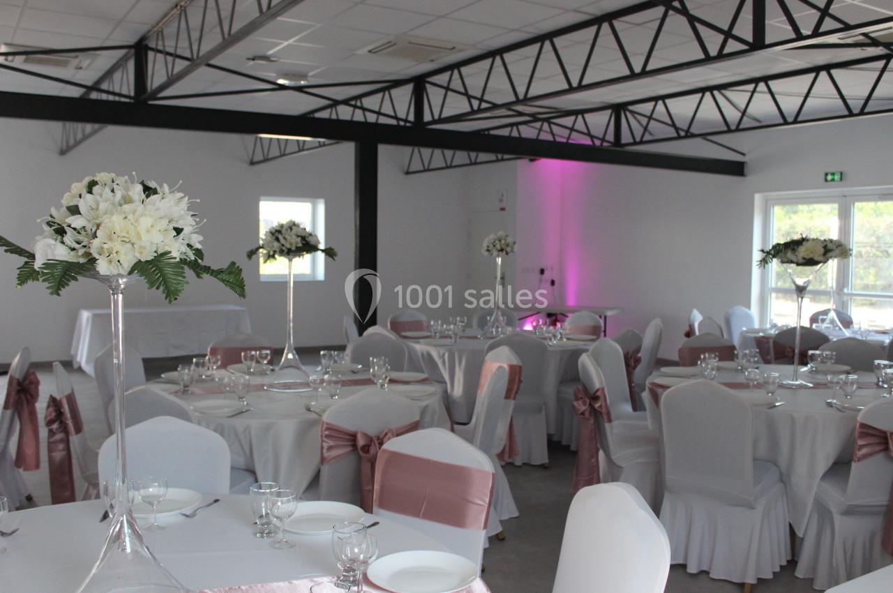Salle de réception décorée avec des tables rondes, nappes blanches, chaises ornées de rubans roses et centres de table…