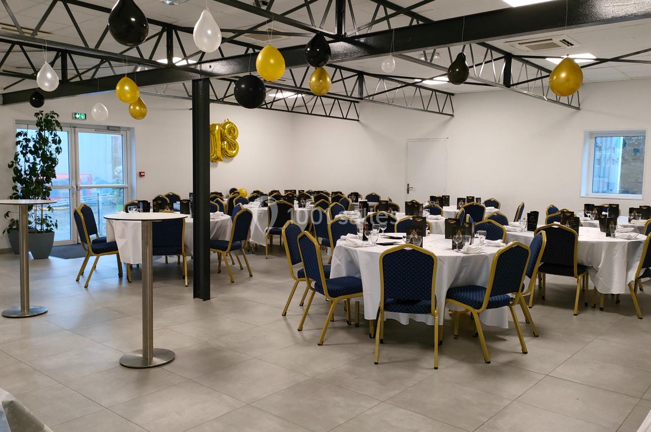 Salle de réception décorée avec des ballons noirs, blancs et dorés, tables rondes dressées avec nappes blanches et chaises…