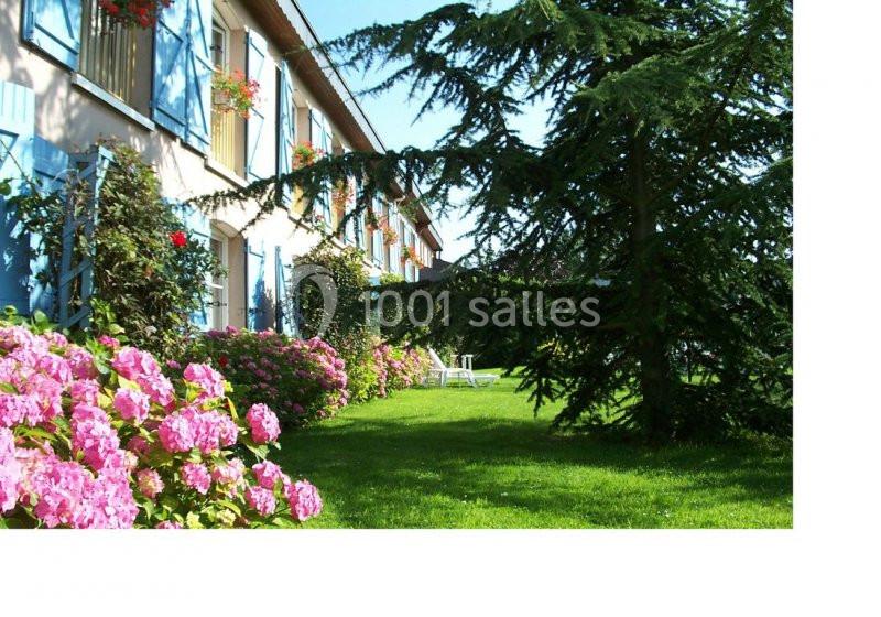 Façade d'une maison avec volets bleus, hortensias roses en premier plan et pelouse bordée d'arbres.