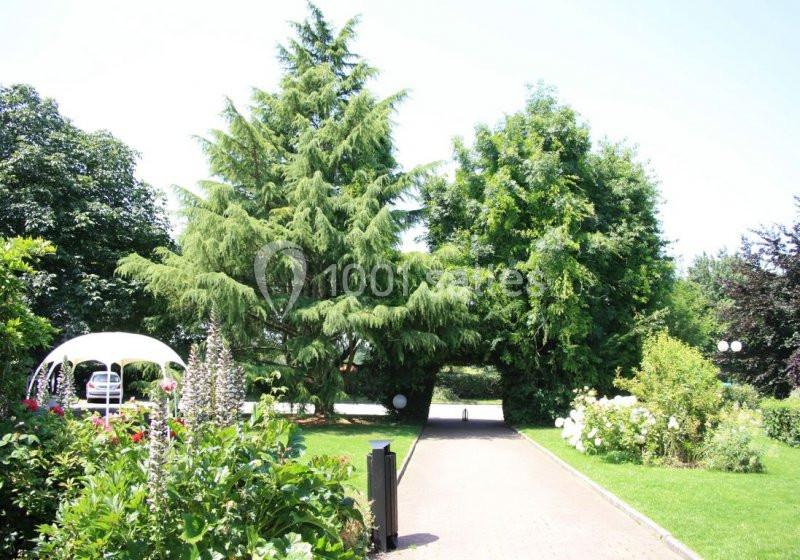 Allée pavée bordée de pelouse, fleurs et arbres, avec une tonnelle blanche visible sur la gauche.