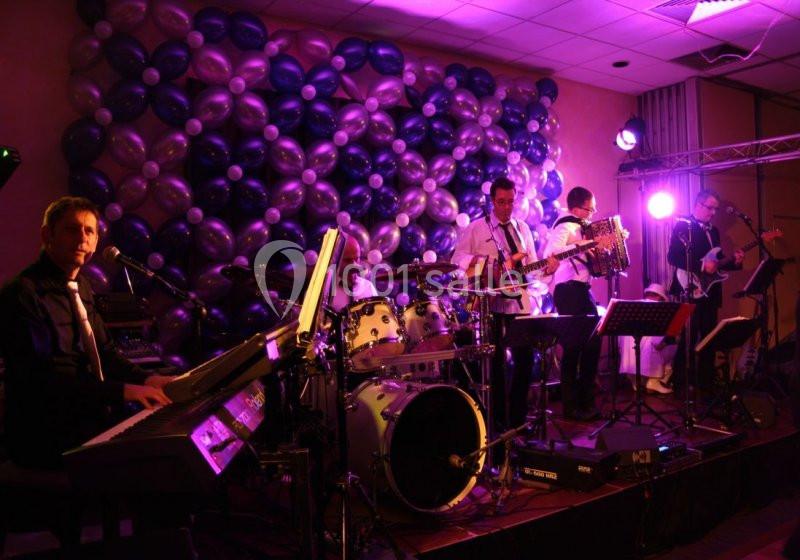 Un groupe de musiciens joue sur scène avec des instruments, devant un mur décoré de ballons colorés.