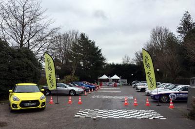 Plusieurs motos stationnées dans un parking entouré d'arbres, avec un véhicule utilitaire en arrière-plan.